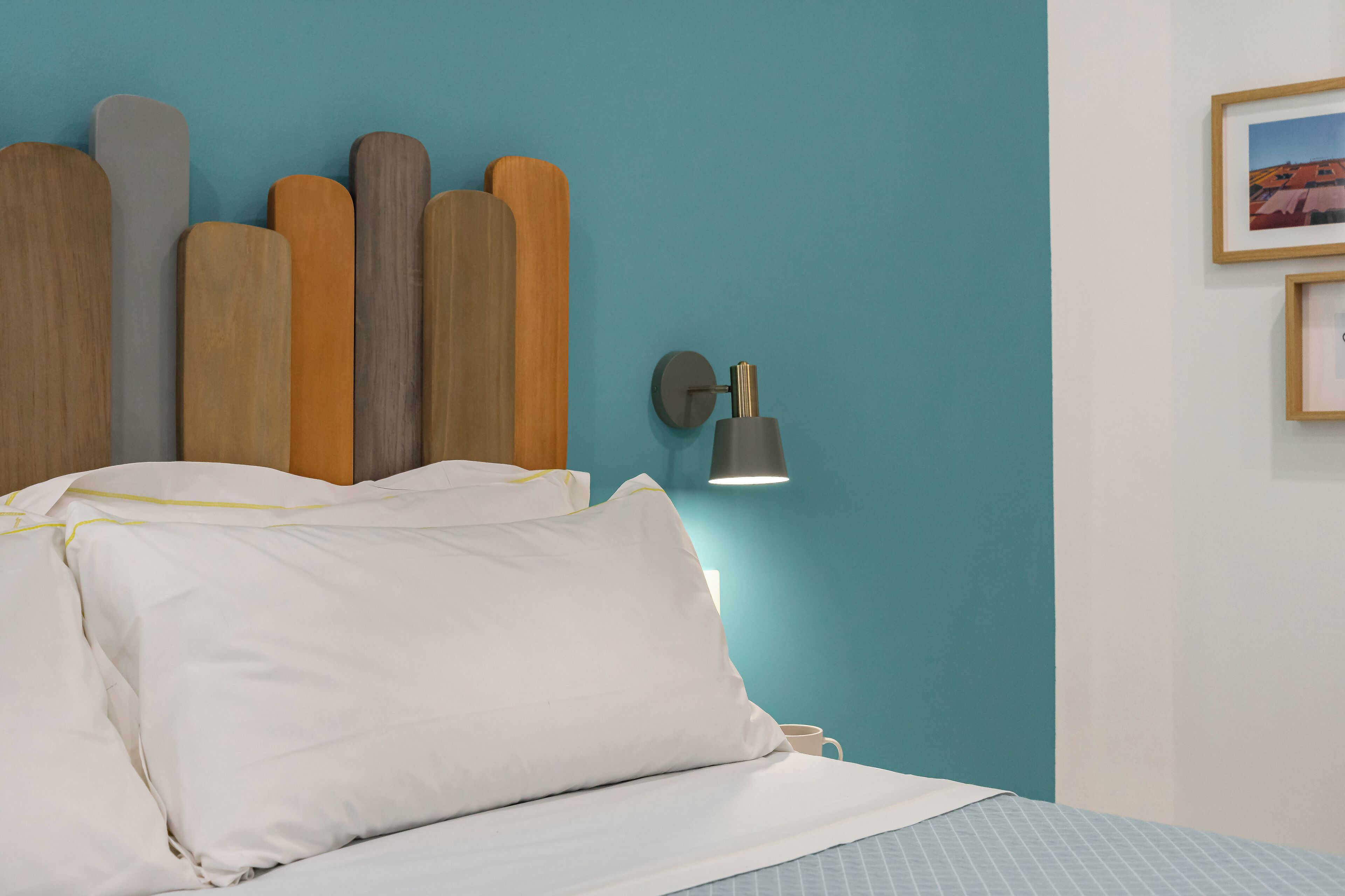 Un lit avec une literie blanche, une lampe fixée au mur et un tableau encadré accroché au mur.