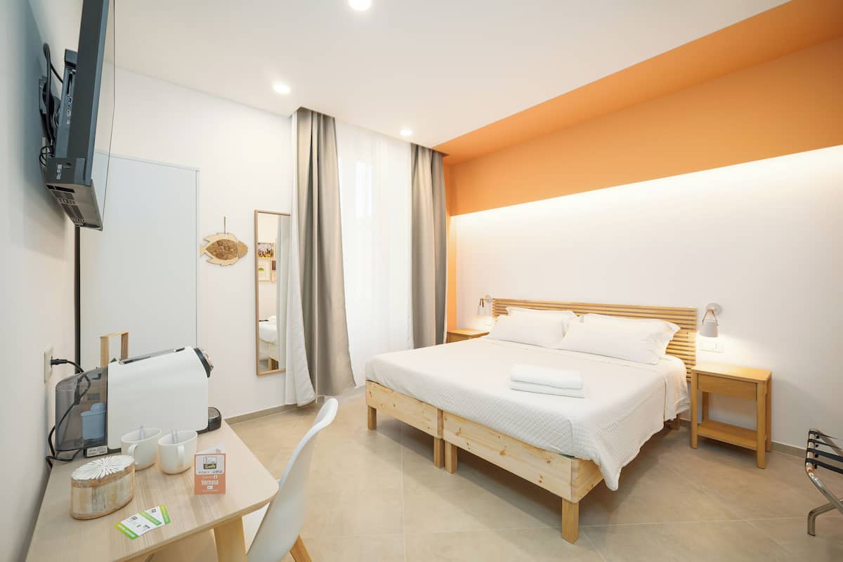 A modern hotel room with a wooden bed, a desk with a computer, a mirror, and a TV mounted on the wall.