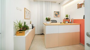 Un área de recepción con mostrador de madera, armarios blancos y naranja, una planta en maceta y un cuadro en la pared.