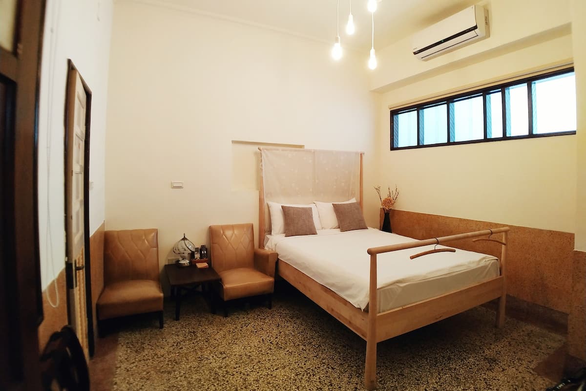 A bedroom with a wooden bed, a small table, two chairs, a window with blinds, and an air conditioning unit.