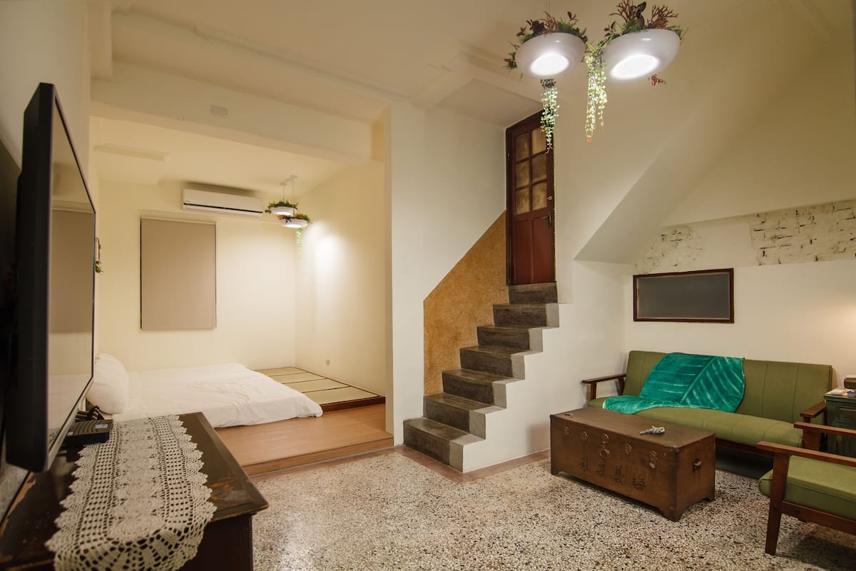 A room with a bed, a desk with a computer, a green sofa, a wooden chest, and a staircase.