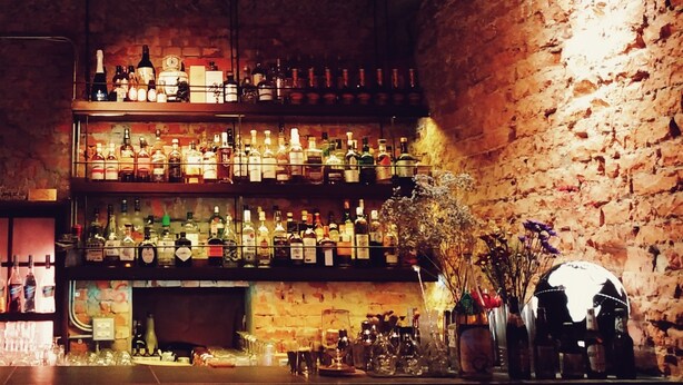 A bar with multiple shelves of bottles and glasses, a neon sign, and a brick wall. - GOIN Old House & Bar (Tainan)