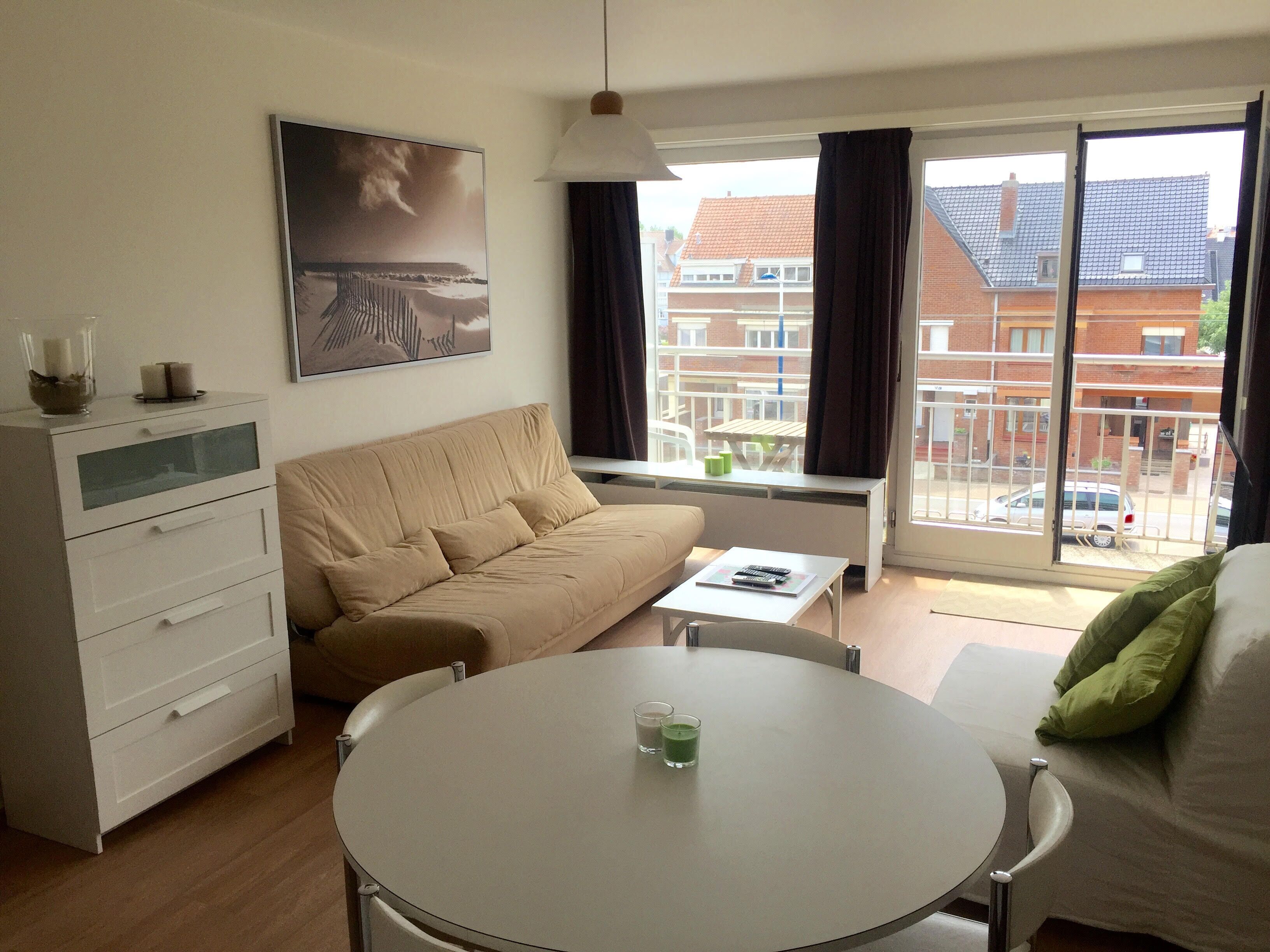 A modern living room with a beige sofa, a white coffee table, and a dining table. There is a large window with curtains, a painting on the wall, and a view of a residential area outside.