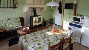Ett mysigt vardagsrum med en soffa, ett matbord med ett mönstrat bordsduksöverdrag, en inbyggd vedkamin, en tv och ett köksområde med mikrovågsugn och kylskåp.