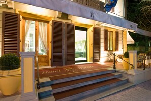 A hotel entrance with a set of stairs leading up to a door, a doormat with the hotel's name, and a flag above the entrance.
