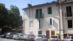 Une rangée de voitures garées le long d’une rue, avec un immeuble doté de balcons et de volets.