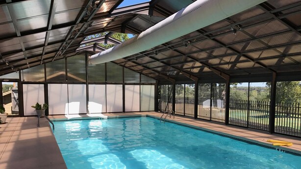 An indoor swimming pool with clear water, surrounded by a covered area with a metal roof and large windows.