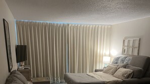 A modern living room with a sofa, a couch, and a bed. There is a TV on a stand, a lamp, and a window with curtains.