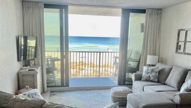 A spacious living room with a large sofa, a smaller couch, and a TV on the wall. The room has a balcony with a view of the beach and ocean.