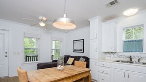 Une cuisine avec des meubles blancs, une table à manger en bois, des chaises en osier et un ventilateur de plafond.