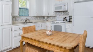 Une cuisine avec des meubles blancs, une table en bois, des chaises en osier, un vase en verre avec des fleurs et une bouilloire.