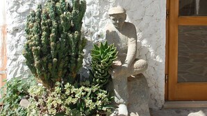 A stone statue of a seated figure next to a cactus plant, with a white textured wall and a wooden door in the background. - Sa Crannaccia (Cabras)