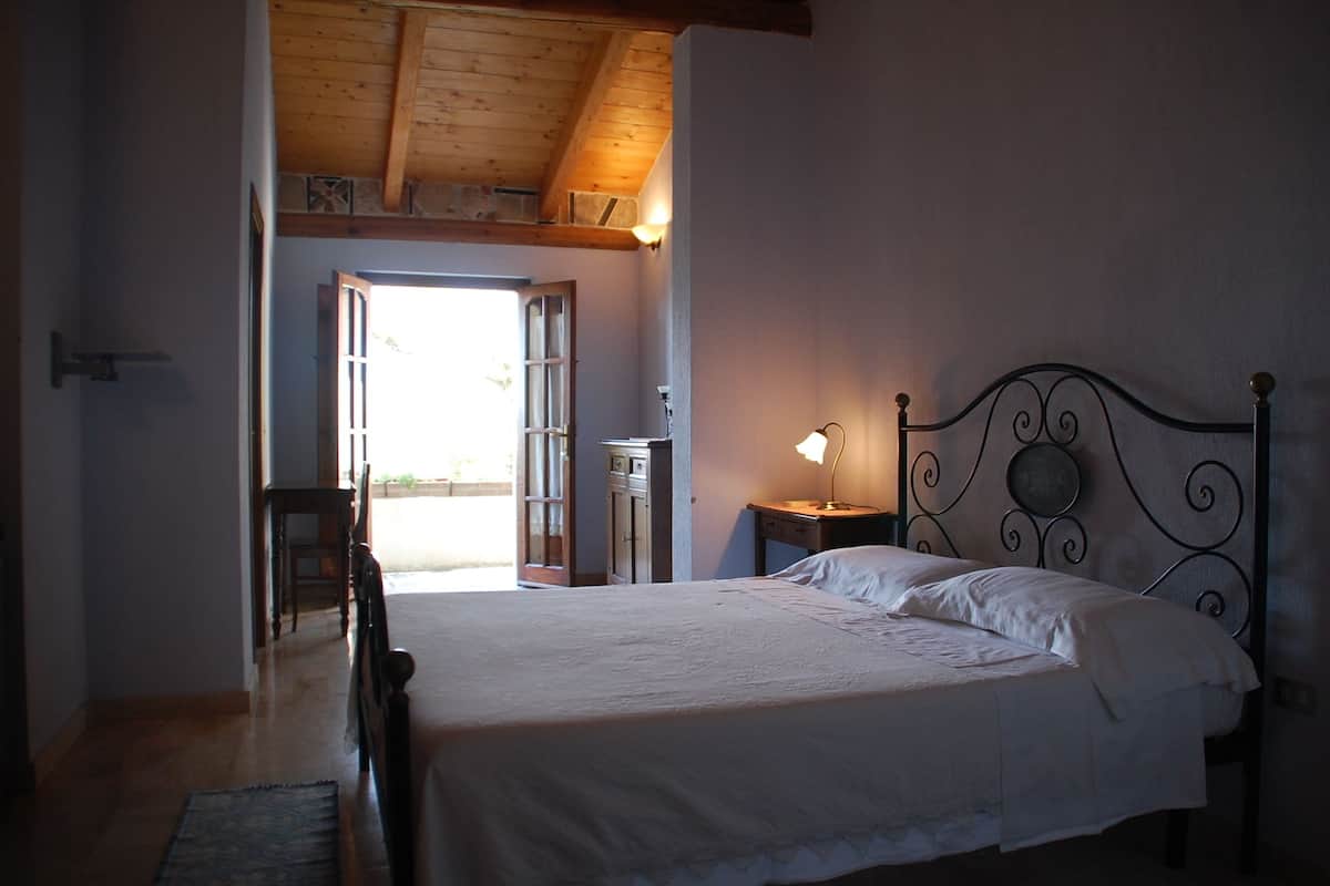A bedroom with a bed, a nightstand, a lamp, and a view of the outside.
