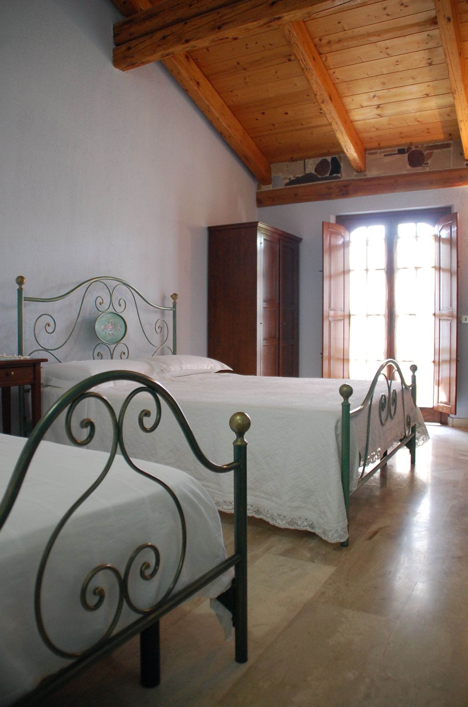 Un dormitorio con dos camas de metal, vigas de techo de madera, un armario de madera y una ventana con contraventanas de madera.