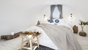 A bedroom with a curved ceiling, a bed with patterned pillows, a bedside table with a candle and a glass of juice, and a wooden chair. - Pnoi Luxury Suites (Santorini)
