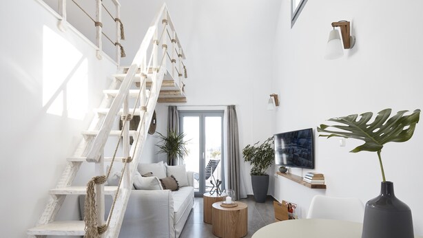 A modern living room with a white staircase, a sofa, a coffee table, a TV, and a potted plant. - Pnoi Luxury Suites (Santorini)