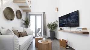 A modern living room with a sofa, coffee table, and TV. There are woven baskets on the wall, a potted plant, and a bookshelf with books. - Pnoi Luxury Suites (Santorini)