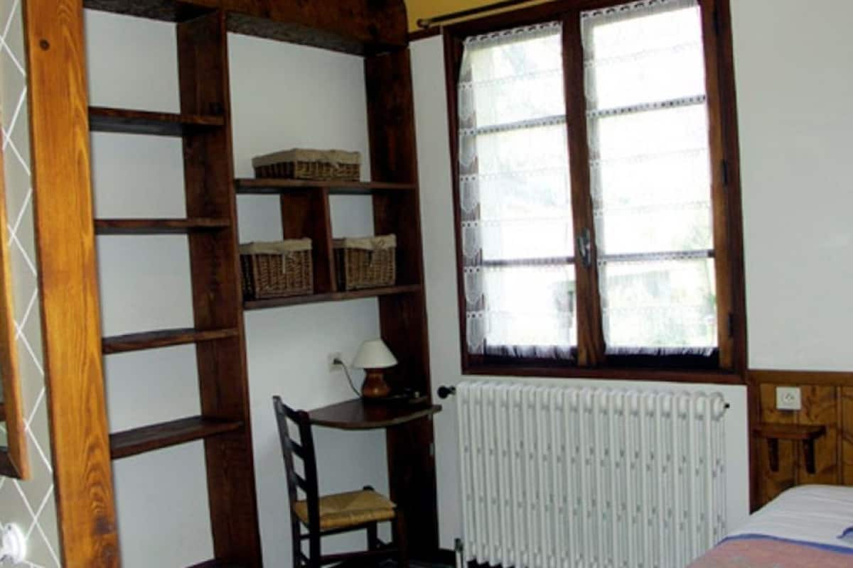 A bedroom with a wooden bookshelf, a chair, a radiator, and a window with lace curtains.
