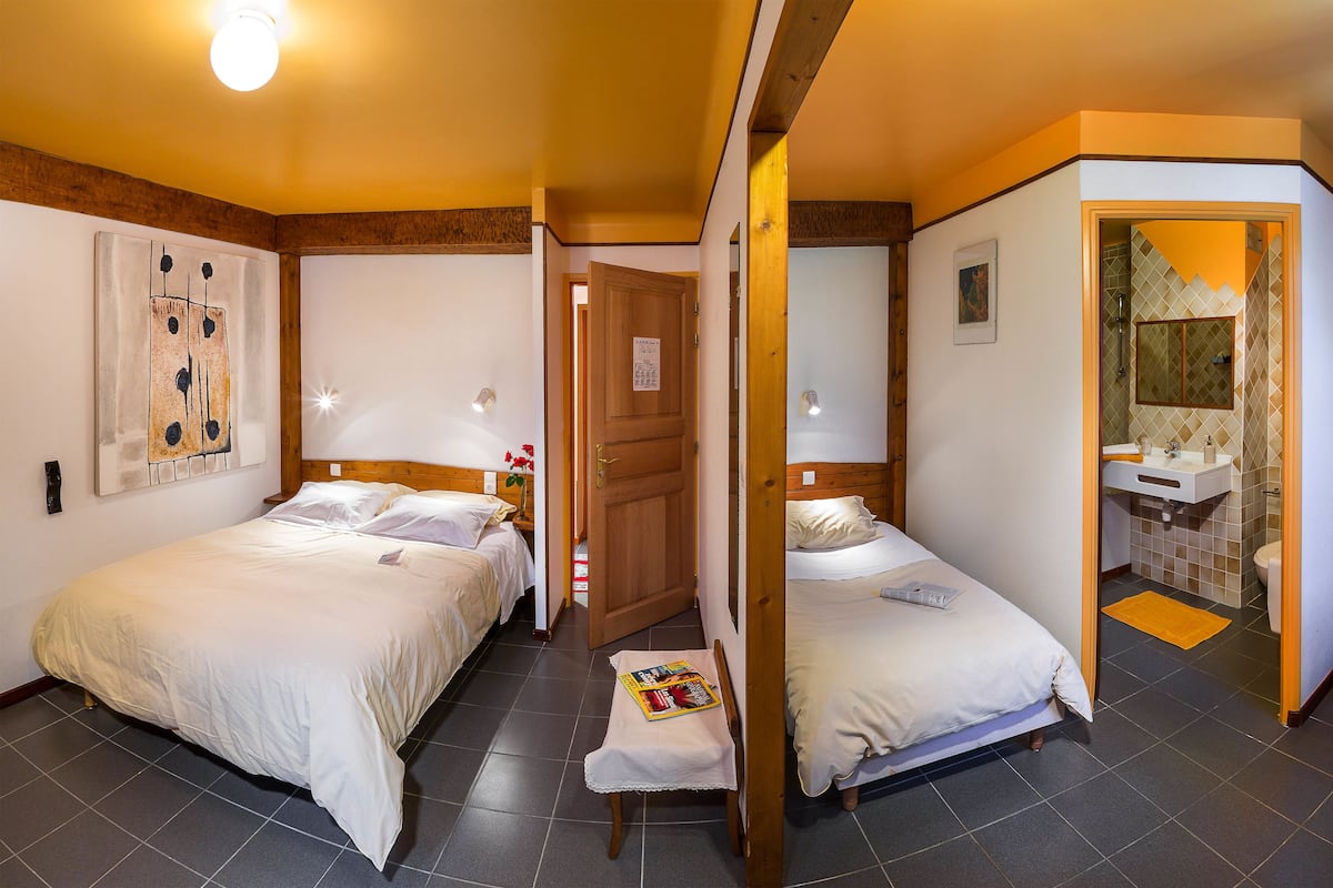 A room with two beds, a wooden door, a small table with a magazine, and a bathroom visible through an open door.