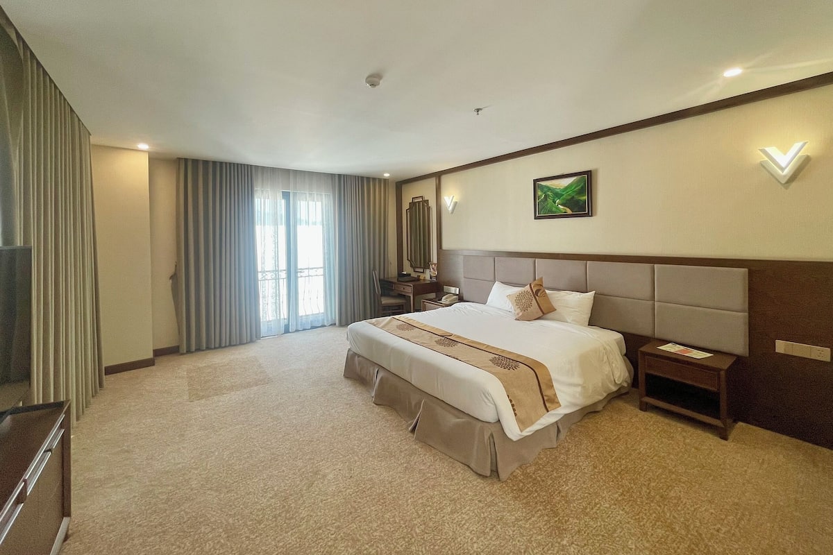 A hotel room with a large bed, a desk, a chair, a nightstand, a mirror, and a window with curtains.