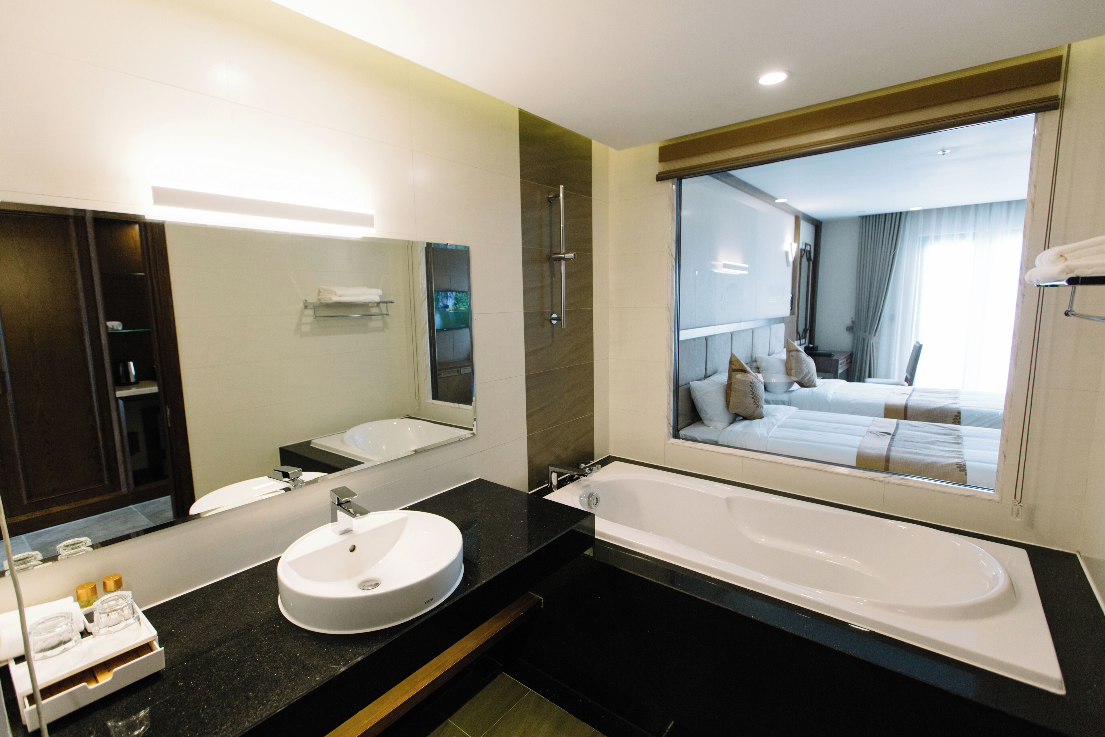 Un baño moderno con un espejo grande, un lavabo redondo, una bañera y vista a un dormitorio a través del espejo.