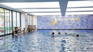 A swimming pool with people in the water and on the edge, a large window, and a wall with 'DUCHUY GRAND HOTEL & SPA' signage.