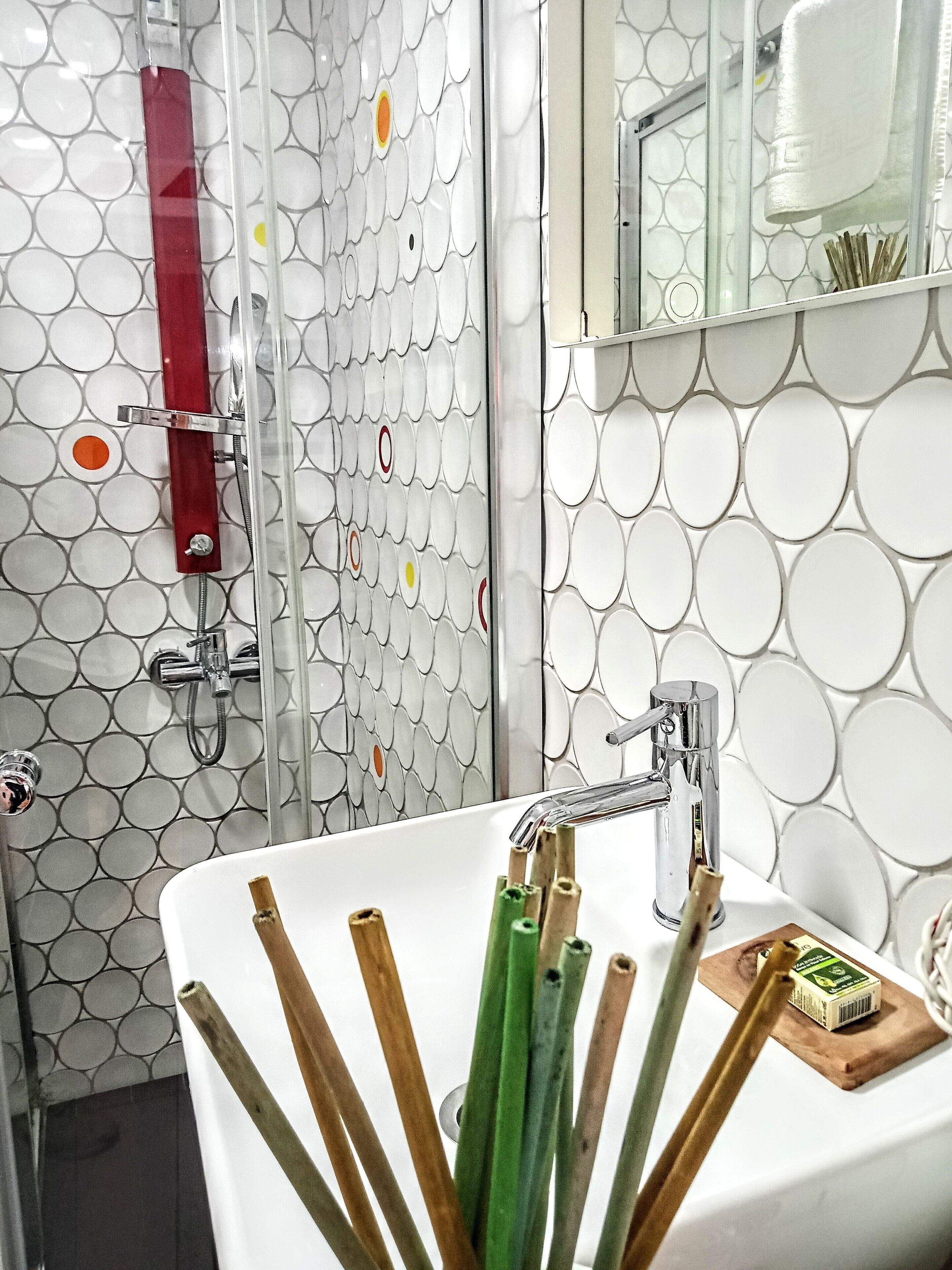 A bathroom with a white sink, chrome faucet, and a mirror above it. There is a shower with a red towel holder and a glass door. The walls have a circular tile pattern.