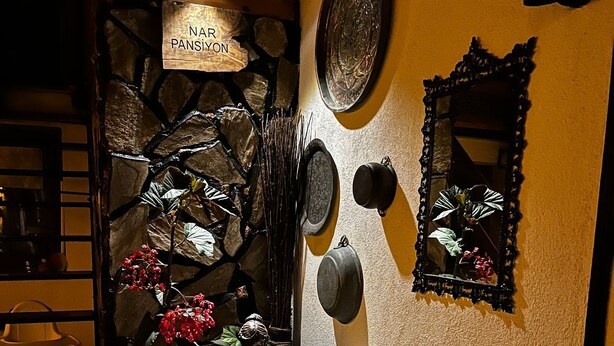 Une pièce chaleureuse avec un mur en pierre, un canapé agrémenté de coussins à motifs et une enseigne suspendue sur laquelle on peut lire « NAR PANSİYON ».