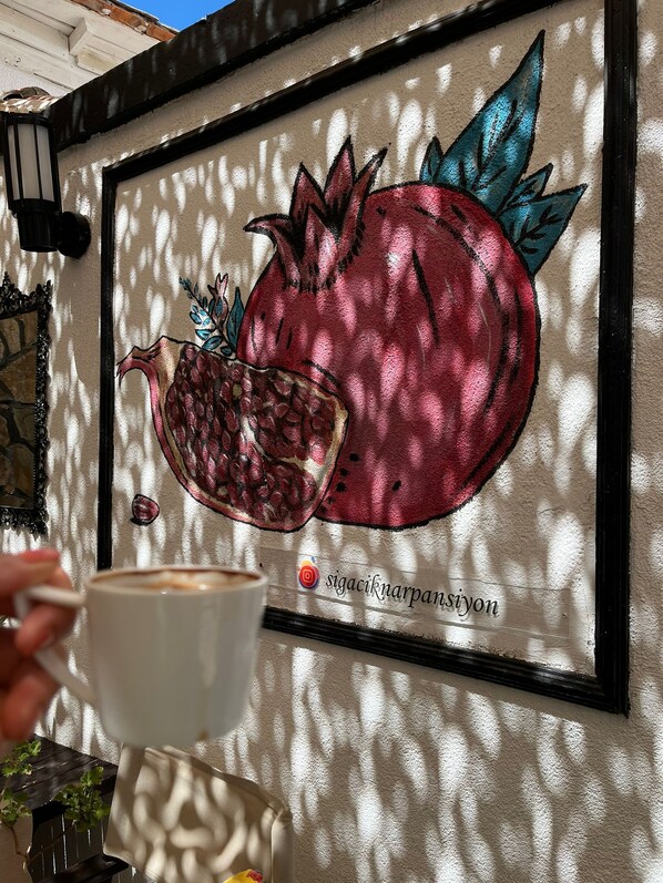 A wall with a pomegranate and pomegranate seeds illustration, a white cup, and a partial view of a plant. - Nar Pansiyon Cafe (Seferihisar)