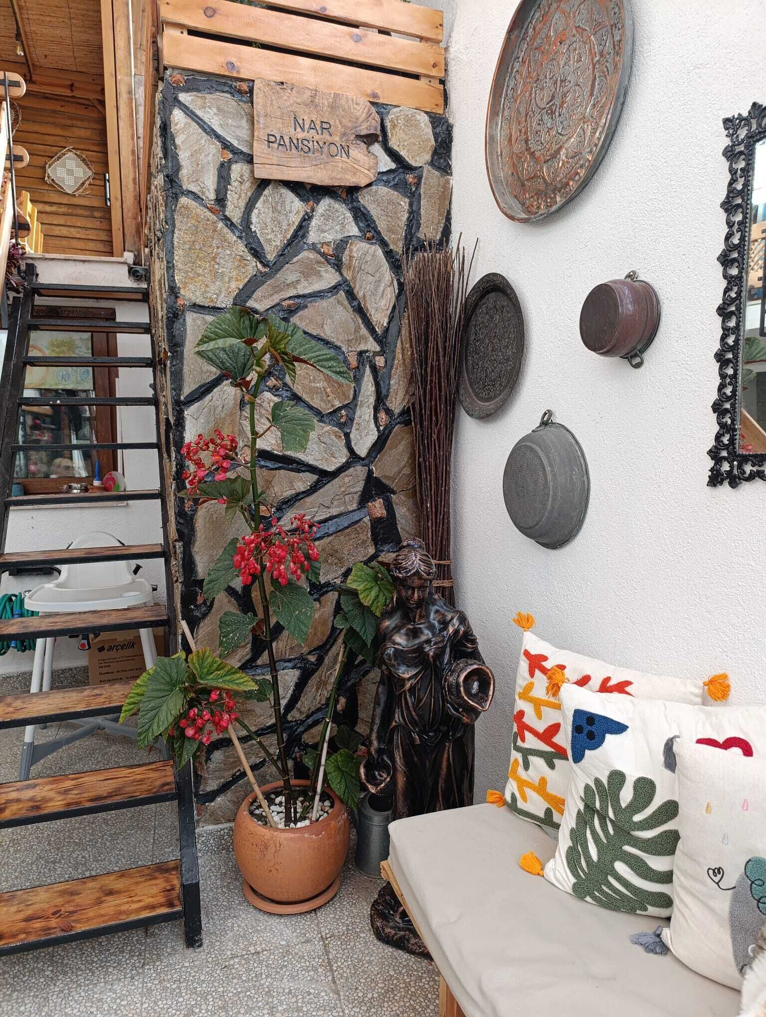 A cozy interior with a stone wall, a wooden sign reading "NAR PANSIYON," and a variety of decorative items including pots, a statue, and colorful cushions.