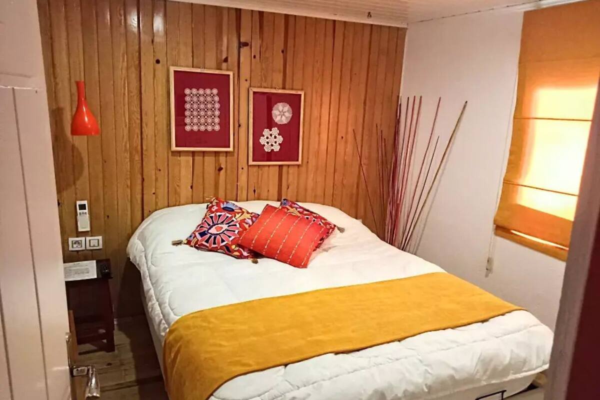 Quarto com parede de madeira, cama com colcha branca e amarela, e duas fotos emolduradas.