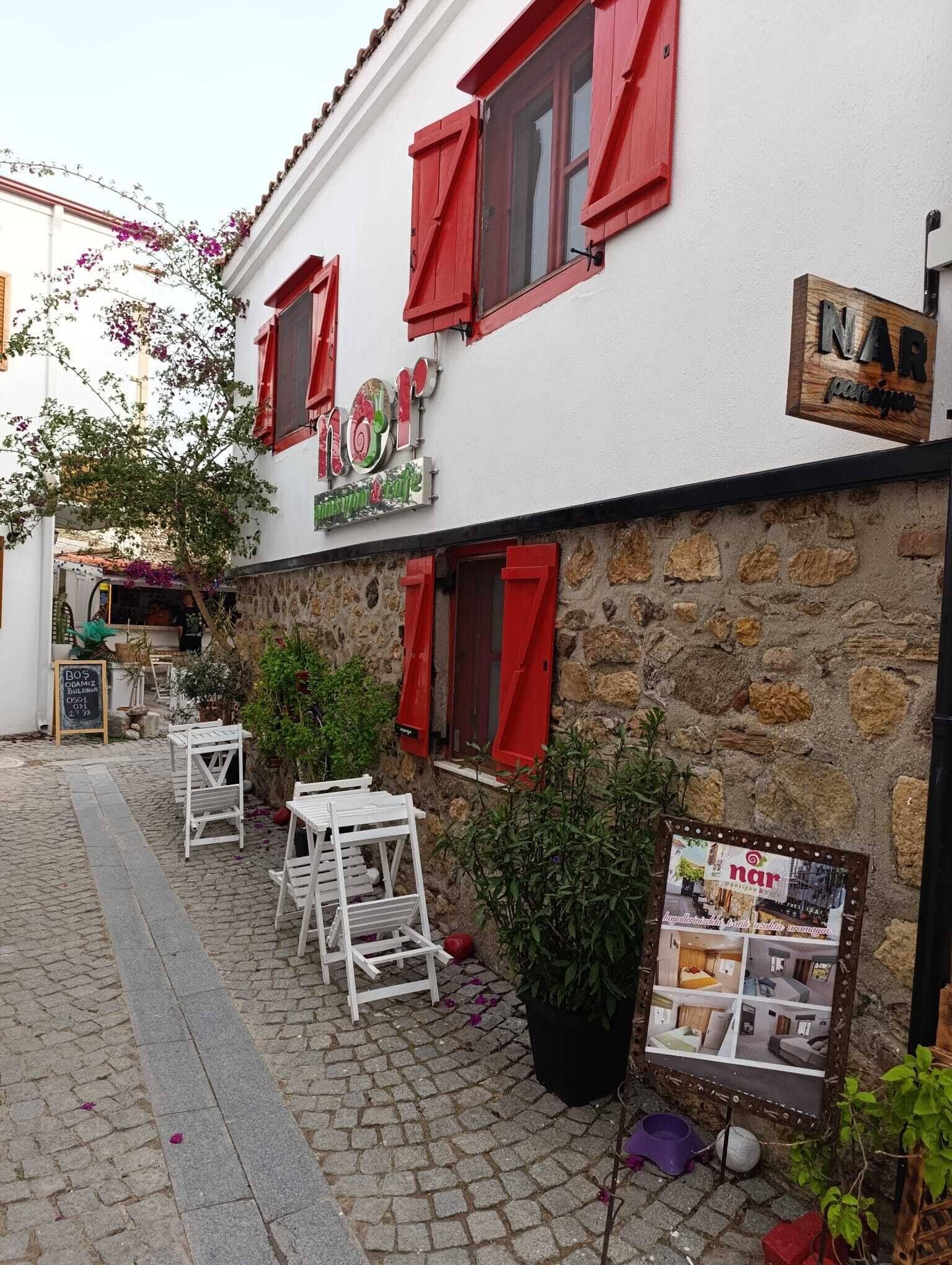 A building with a sign reading "NAR" and a stone wall. There are outdoor tables and chairs, and a signboard with images of rooms.