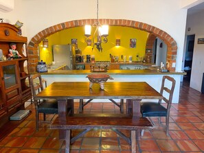 Uma área de refeições com mesa e cadeiras de madeira, um arco de tijolos e uma cozinha ao fundo.