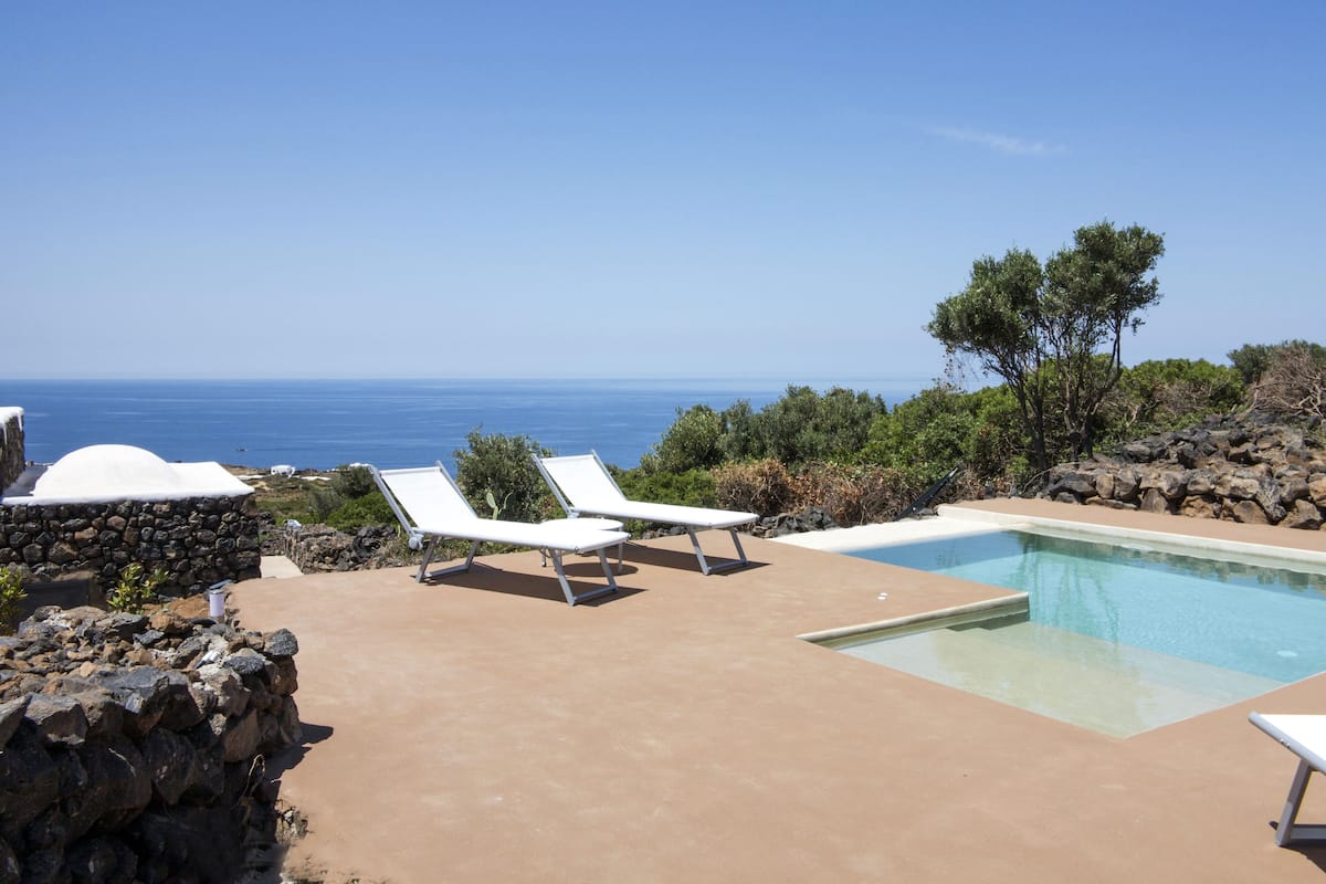 A poolside area with two white lounge chairs, a stone wall, and a view of the ocean.