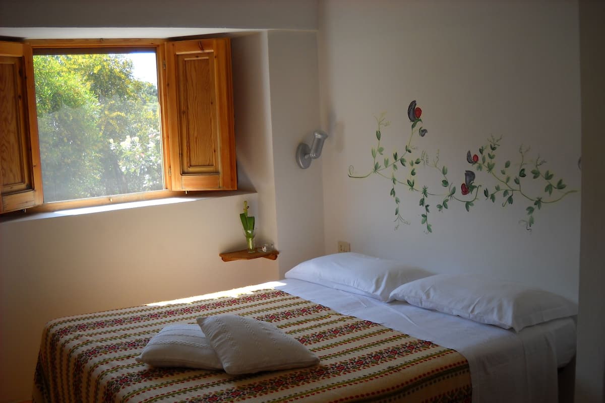 A bedroom with a bed, a window with wooden shutters, a wall-mounted shelf, and a floral wall decal.
