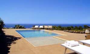 A pool with a clear view of the ocean, lounge chairs, and a table with towels. - Dammusi Sciuvechi Resort (Pantelleria)