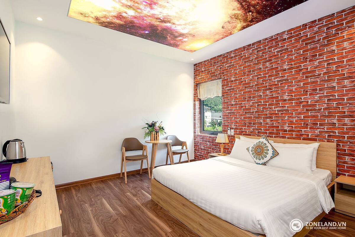 Una habitación de hotel moderna con pared de ladrillo, una cama de madera, un escritorio pequeño con un hervidor y un mural en el techo.