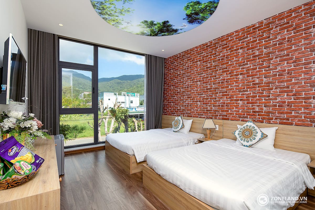 Habitación de hotel con dos camas, una pared de ladrillo y un ventanal con vistas a zonas verdes y un edificio.