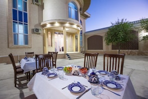Un área de comedor al aire libre con mesas dispuestas para dos, con vajilla azul y blanca, y un edificio con estilo arquitectónico clásico al fondo.