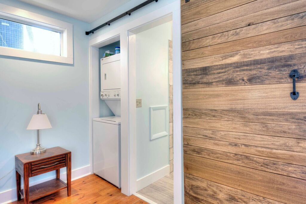 Una habitación pequeña con piso de madera, una puerta blanca, una mesita con lámpara y una ventana.