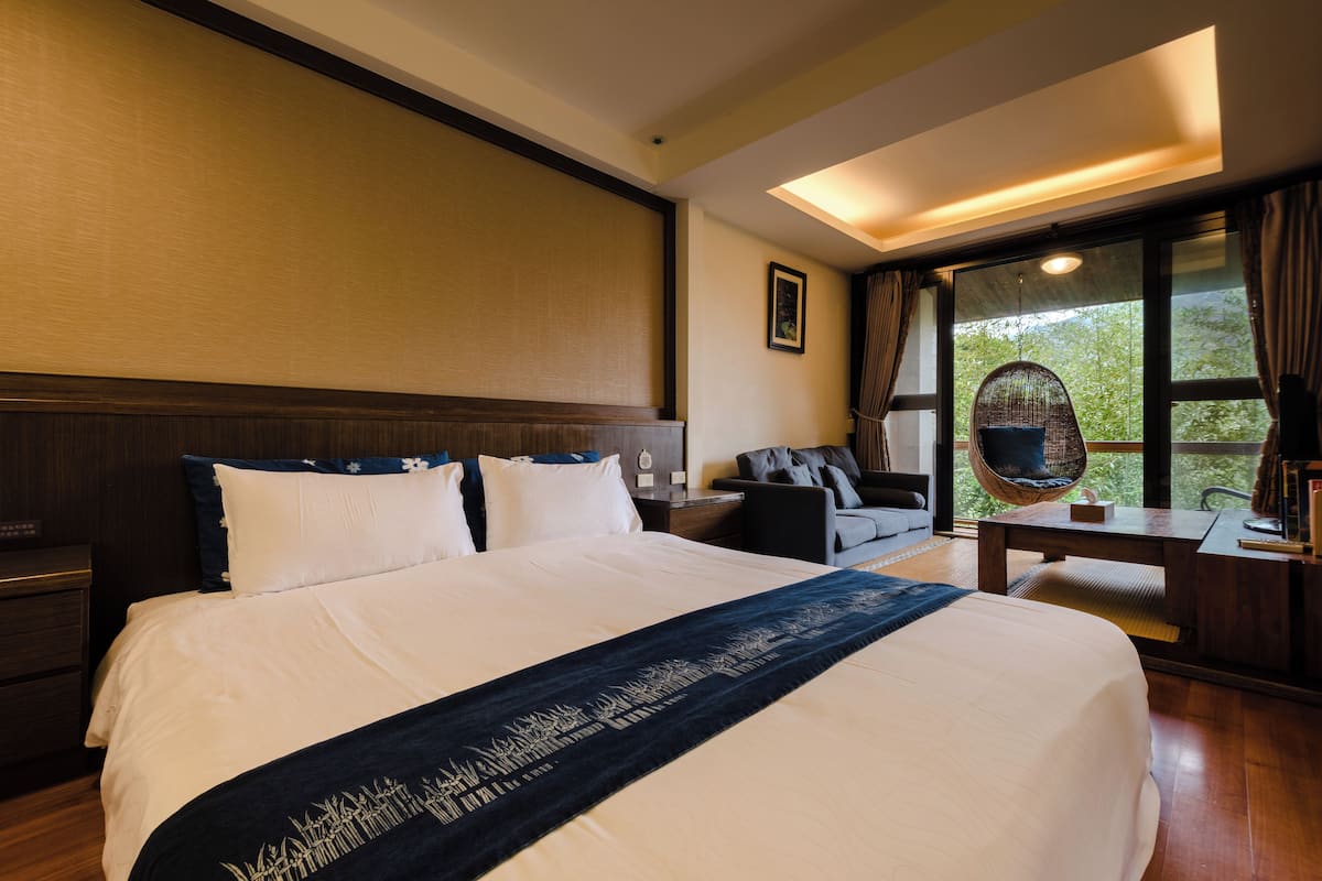A hotel room with a large bed, a desk, a chair, and a view of greenery outside.