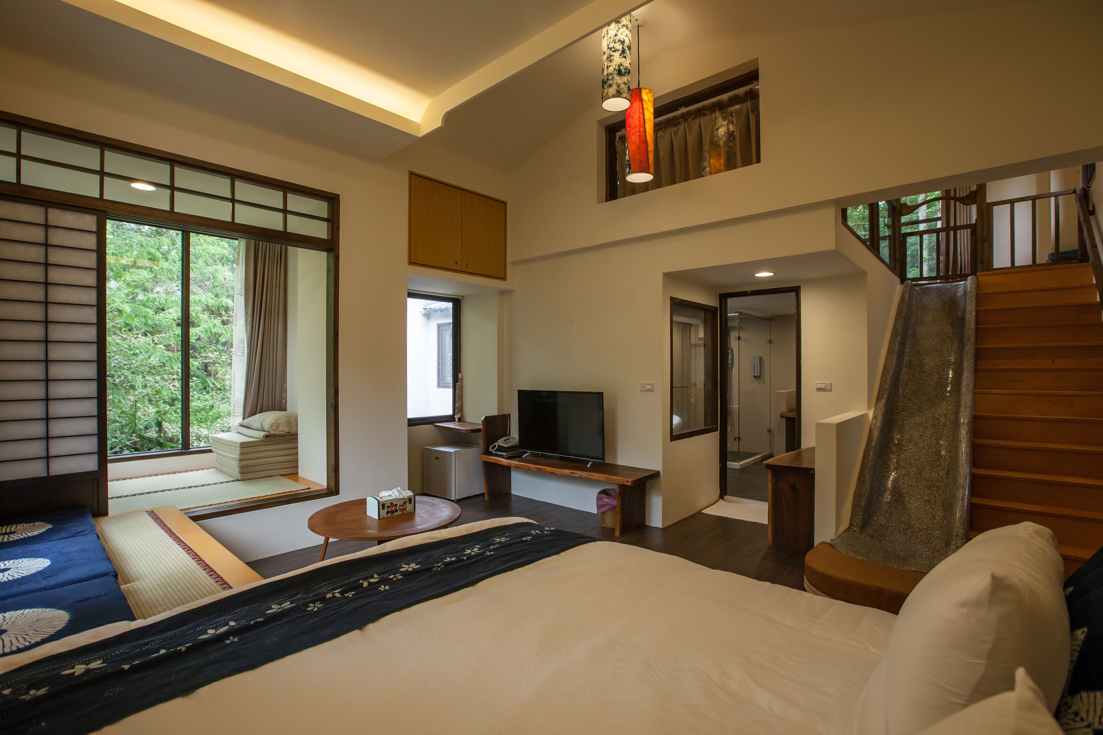 A modern hotel room with a large bed, a TV, a desk, and a view of greenery outside.