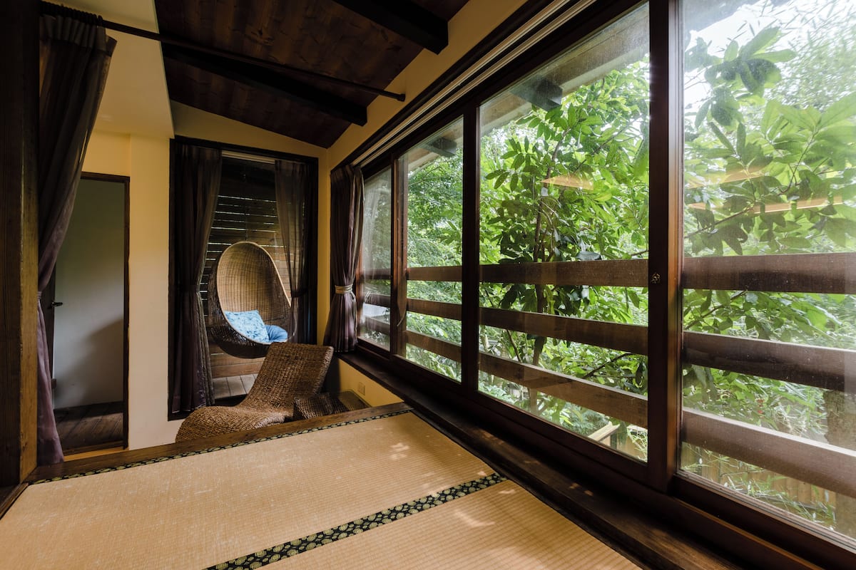 A traditional Japanese room with tatami flooring, a low wooden bed, and a view of lush greenery through large windows.