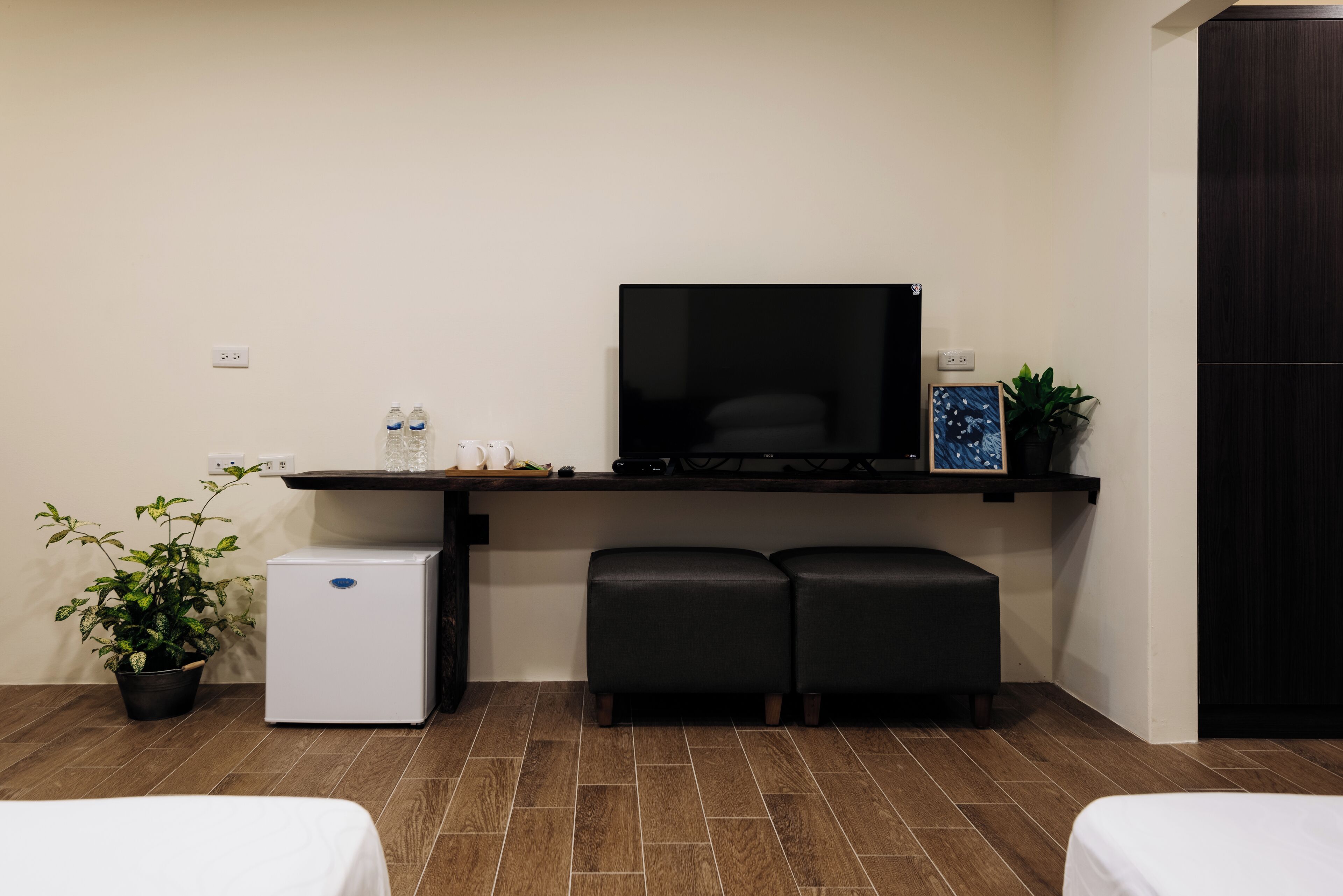 Una habitación pequeña con cama, un refrigerador, un televisor y un banco.