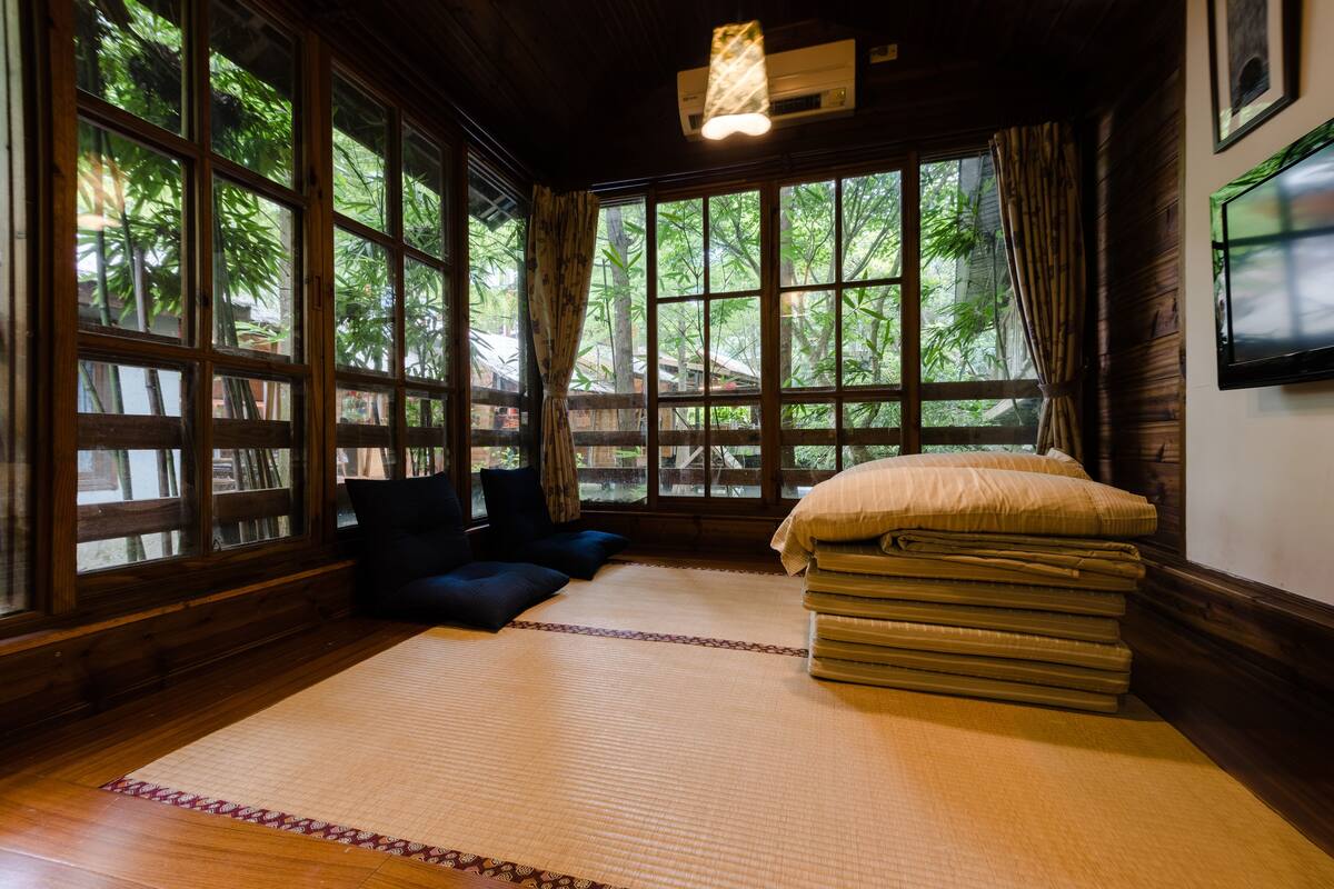 A traditional Japanese room with tatami flooring, a futon bed, and large windows with curtains.