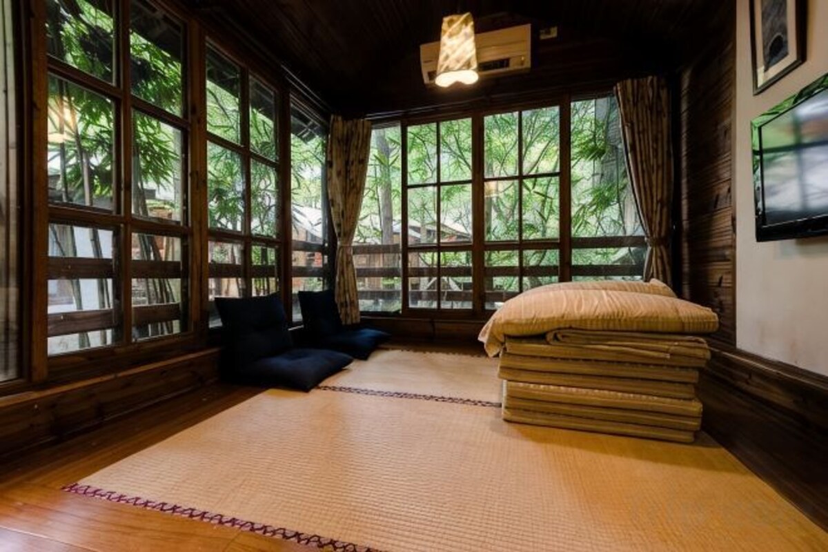 A room with tatami flooring, a bed with a folded blanket, a pillow, and a window with a view of greenery.