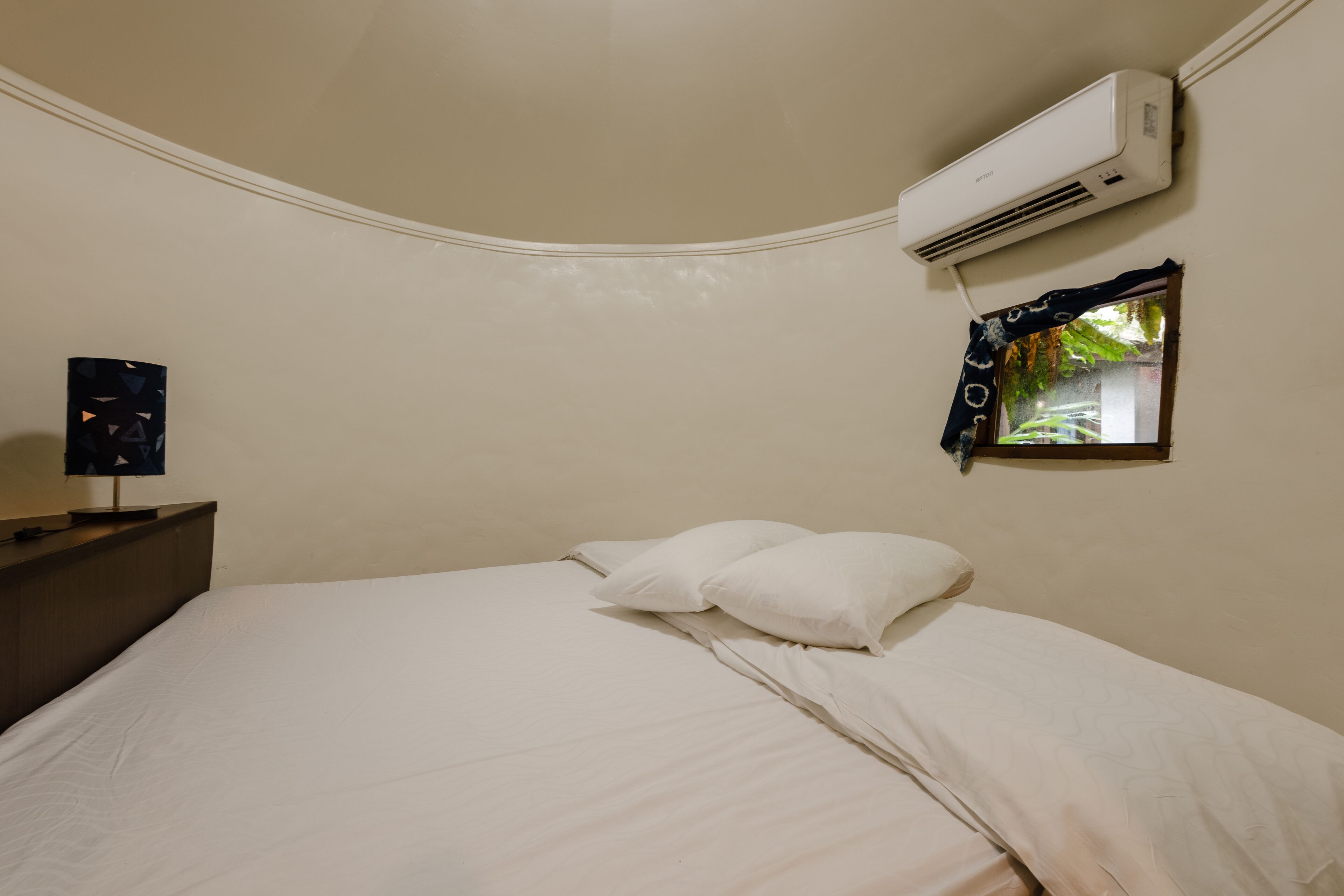 Un dormitorio con cama, almohadas blancas, mesita de noche, aire acondicionado y ventana con vistas al verdor.