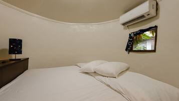 Un dormitorio con cama, almohadas blancas, mesita de noche, aire acondicionado y ventana con vistas al verdor.