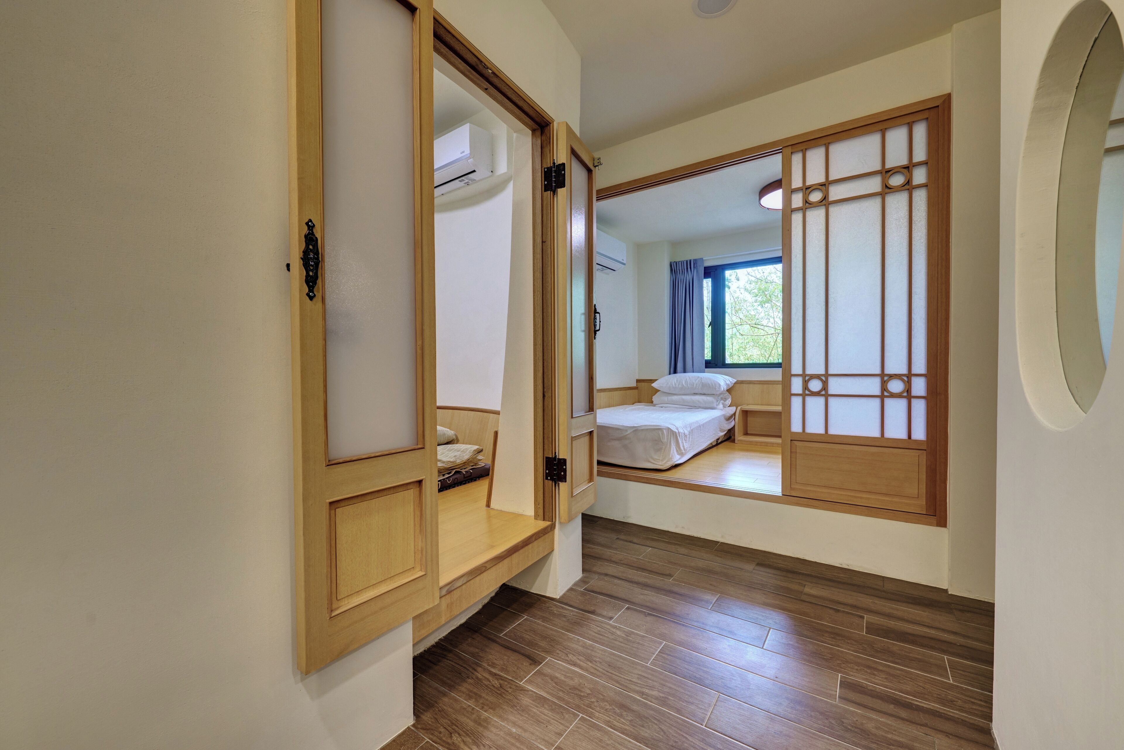 A room with a wooden wardrobe, a bed with white bedding, and a view of the outdoors through a window with a curtain.