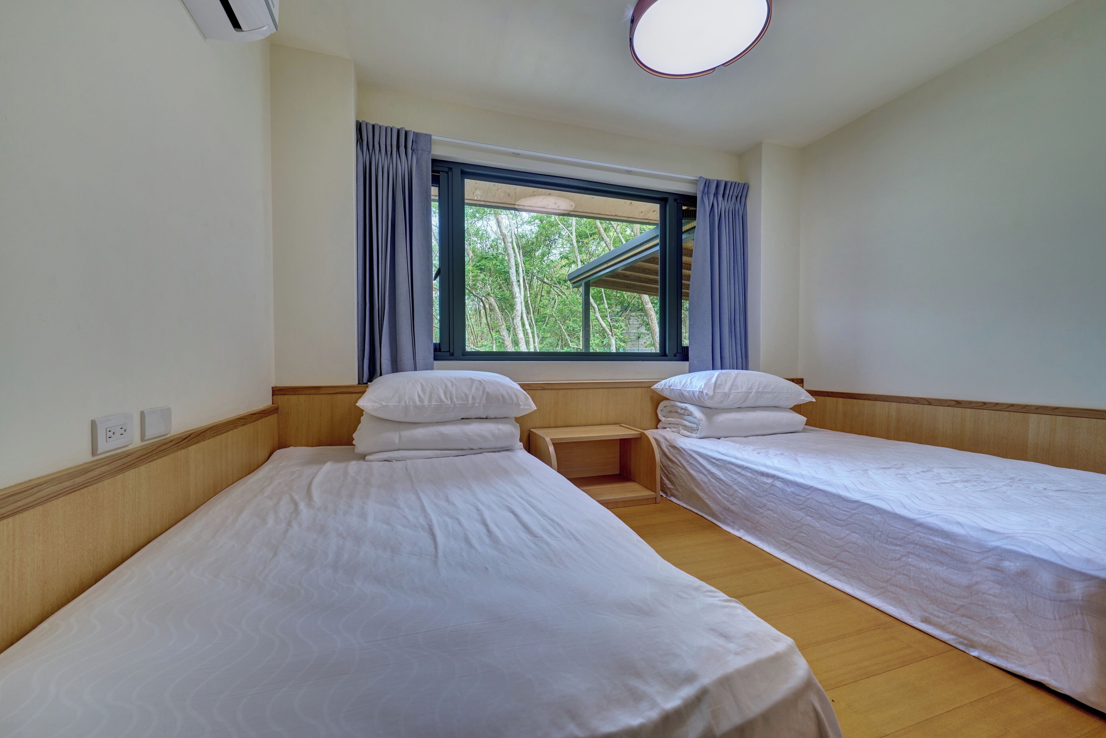 A room with two single beds, a window with curtains, a ceiling light, and a wooden floor.